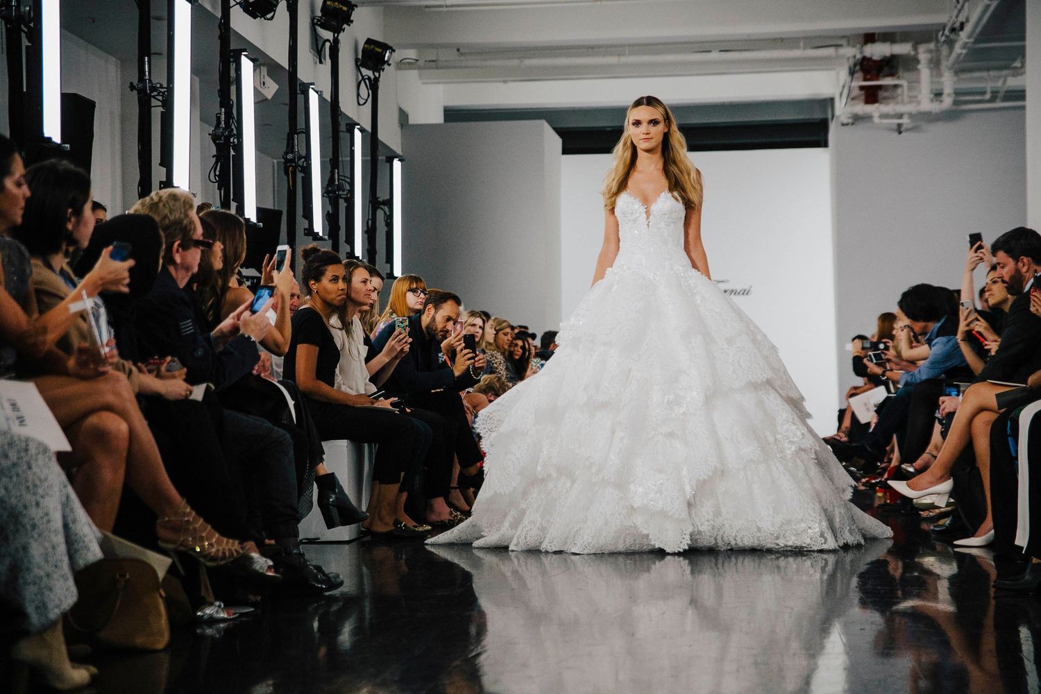 Model walks down a fashion show runway wearing a dramatic white wedding gown with strapless bodice and tulle skirt.