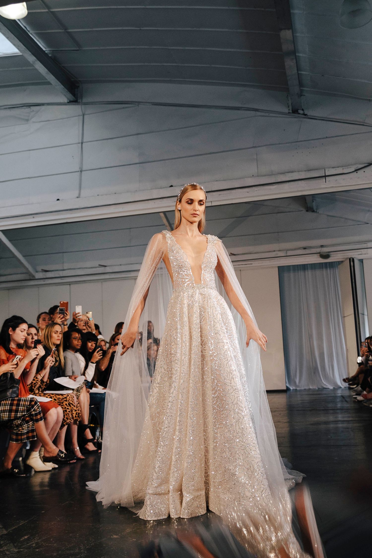 Model walks runway in stunning sparkly white wedding gown with sheer cape details during fashion show.