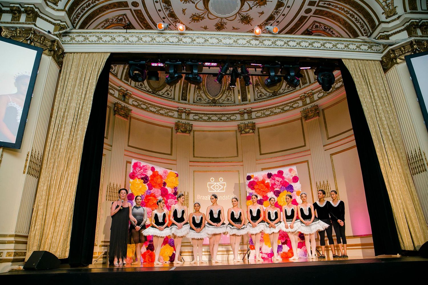 Theater performers on stage in an ornate concert hall with elegant architecture and decorative ceiling details.