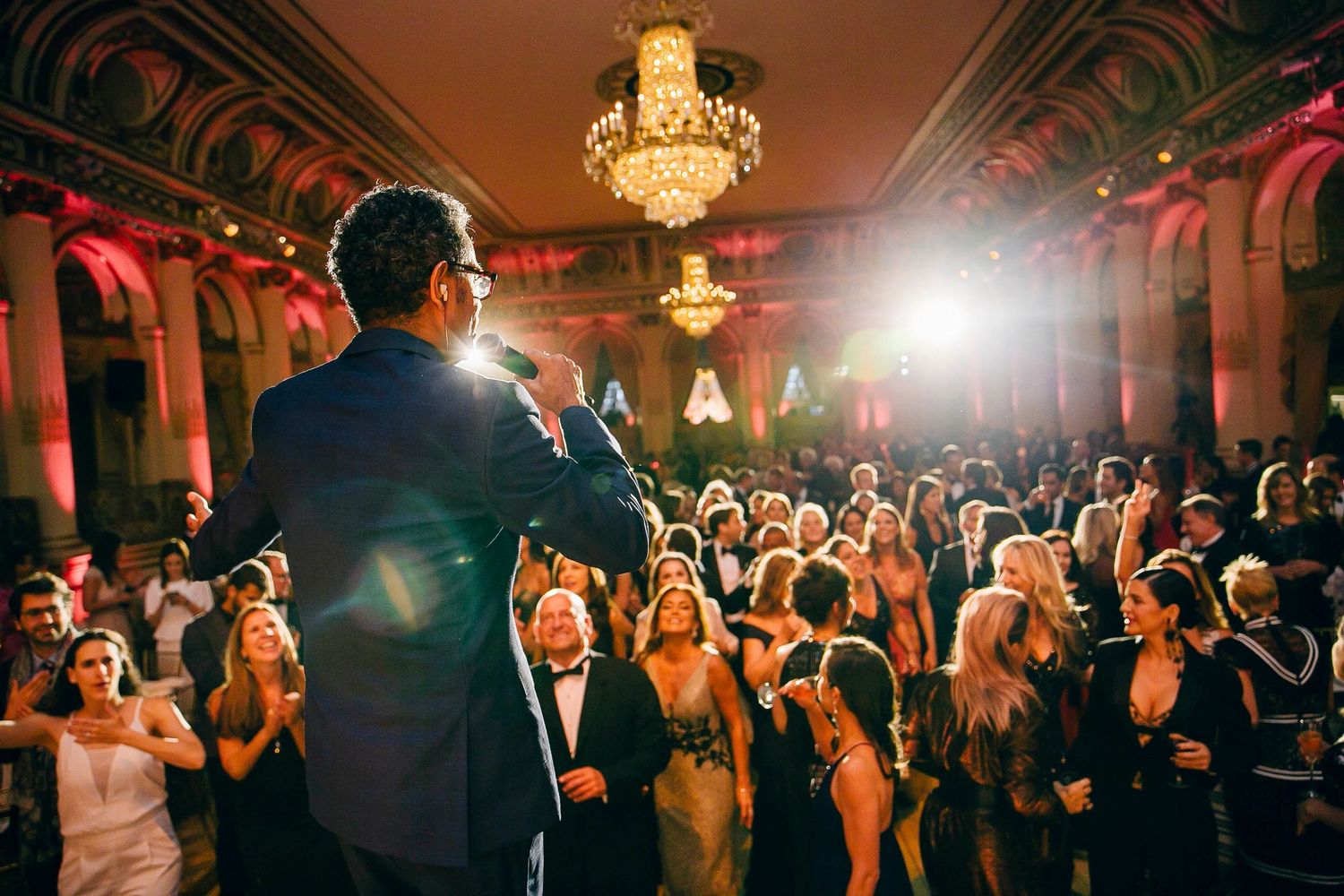 Wedding guests dance and celebrate beneath ornate chandeliers during an elegant evening reception celebration.
