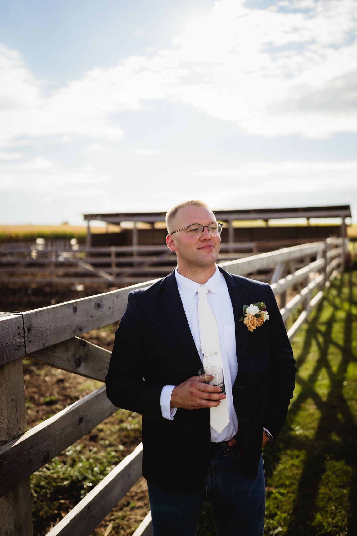 A groom in a black tuxedo and white bowtie stands in front of a wooden fence at sunset with a boutonniere.