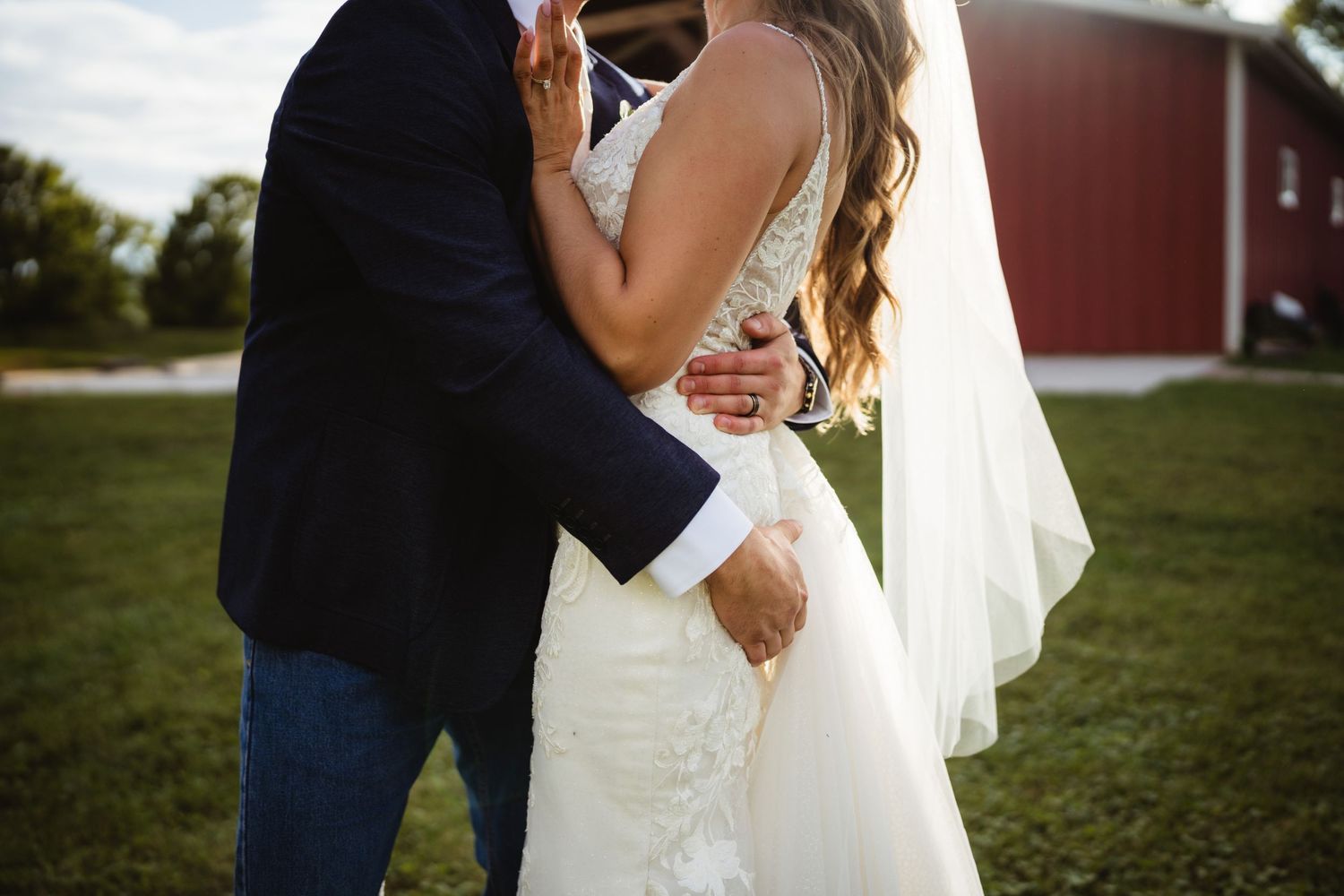 A bride and groom embrace closely during their outdoor wedding at sunset in a grassy field.