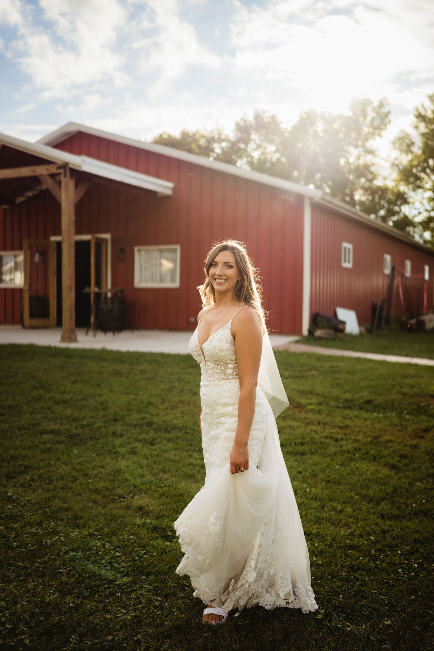 A bride in a lace wedding dress walking along a rustic red barn on a grassy hillside at sunset.