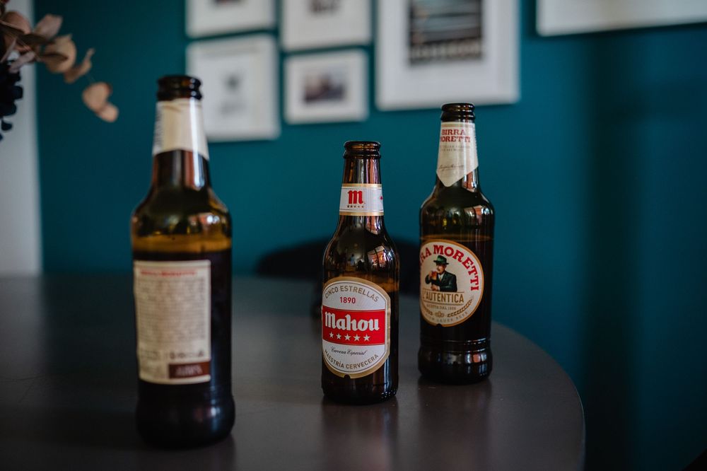 A collection of assorted beer bottles displayed in a row against a dark blue wall with framed artwork.
