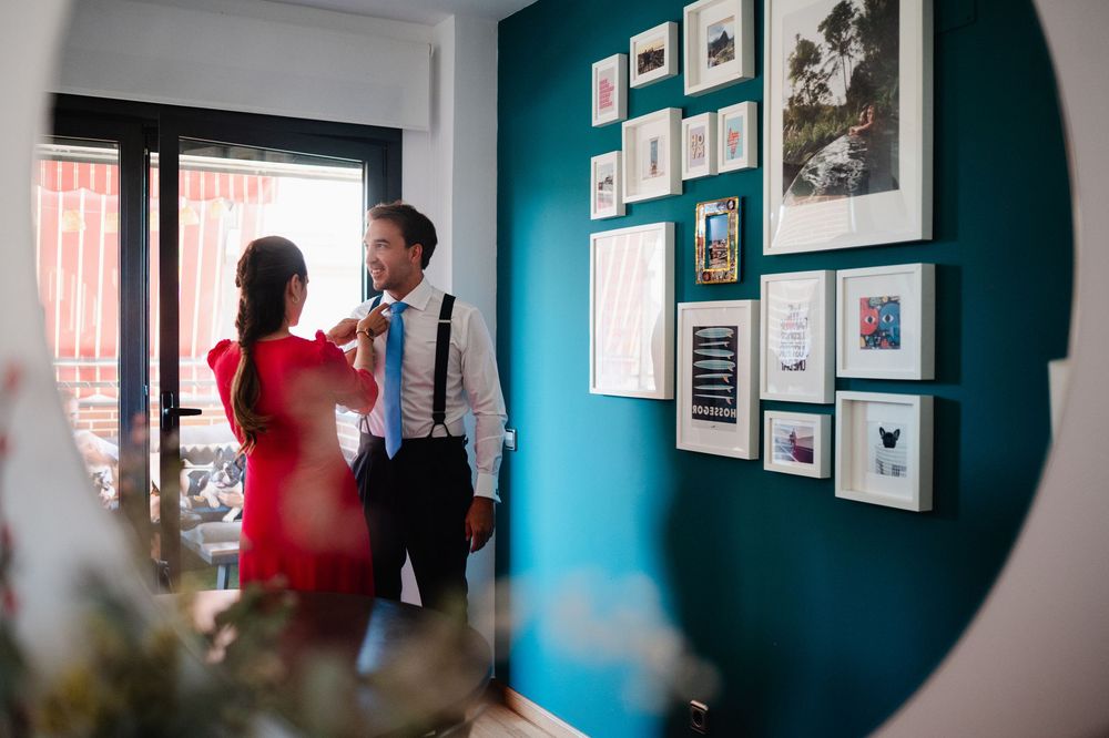 Art pieces hang on turquoise walls in a gallery style display with viewers examining the works in natural light.