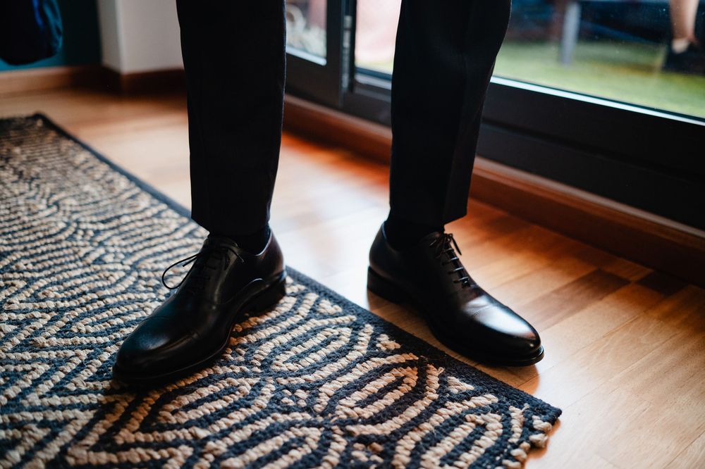 Black dress shoes on a geometric patterned area rug near a window creating shadows on hardwood floor.