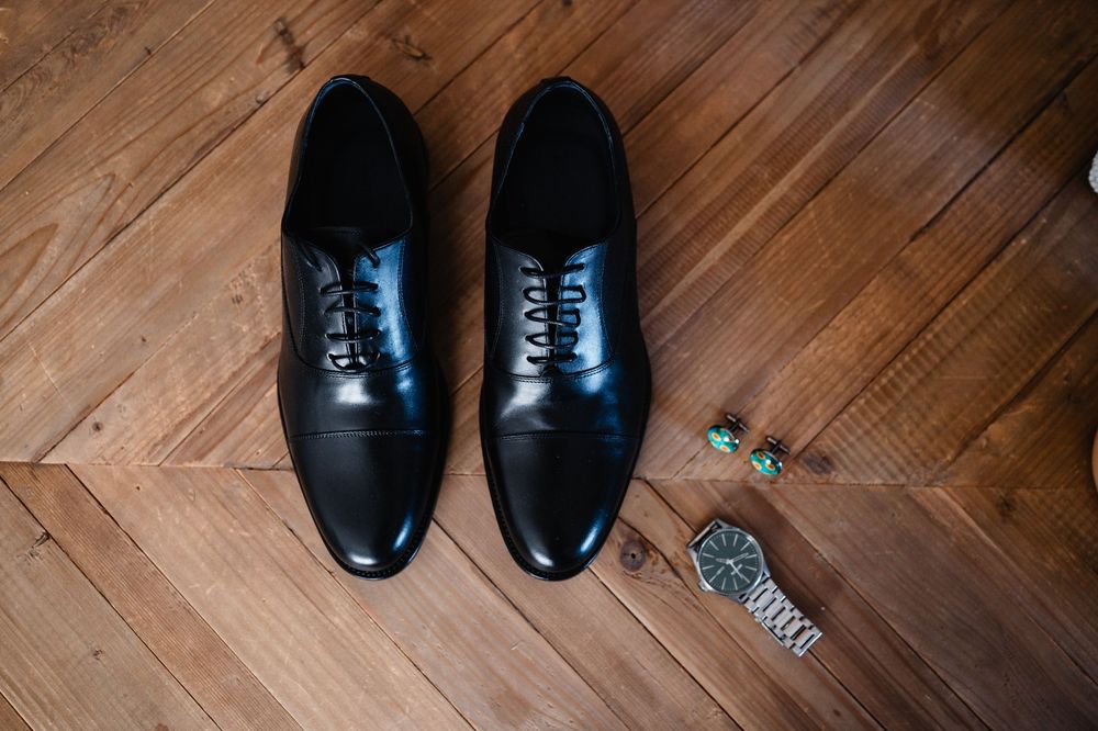 Black formal leather dress shoes arranged in sequence on herringbone wood flooring with watch accessories.