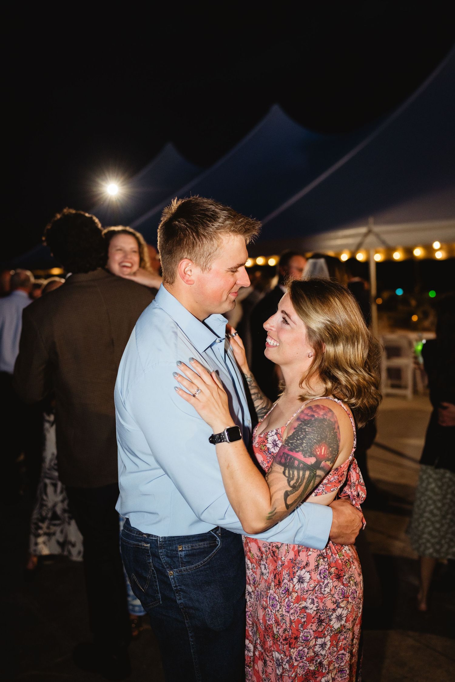 A couple dances together at an outdoor evening event under a tent with string lights glowing in the background.