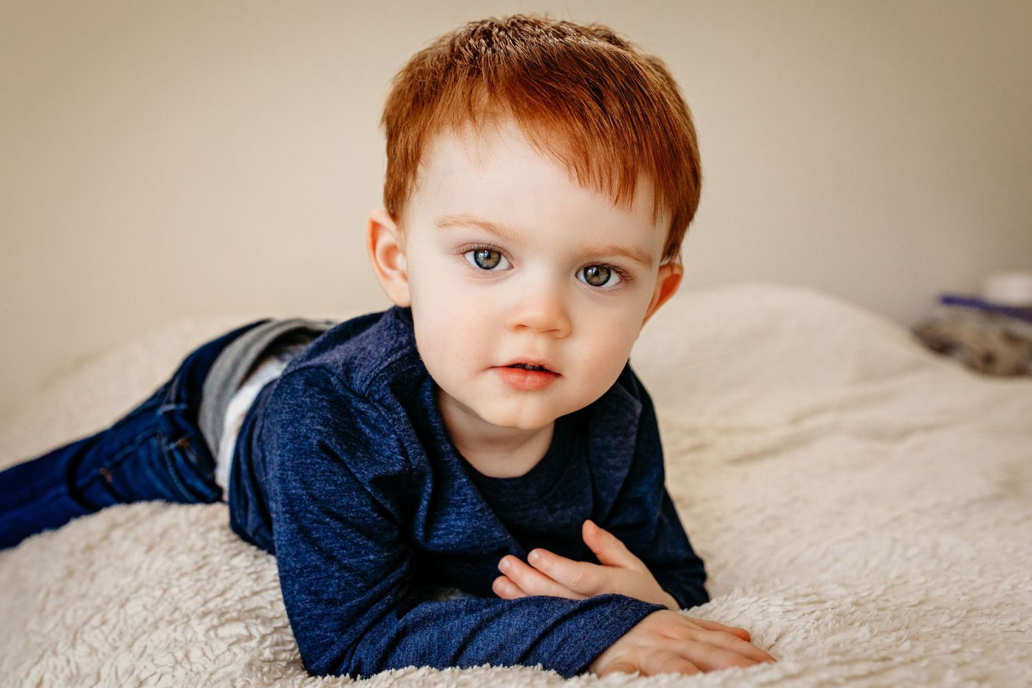 Small child in navy blue outfit lies on cream colored bedding while looking at camera during indoor photo session.