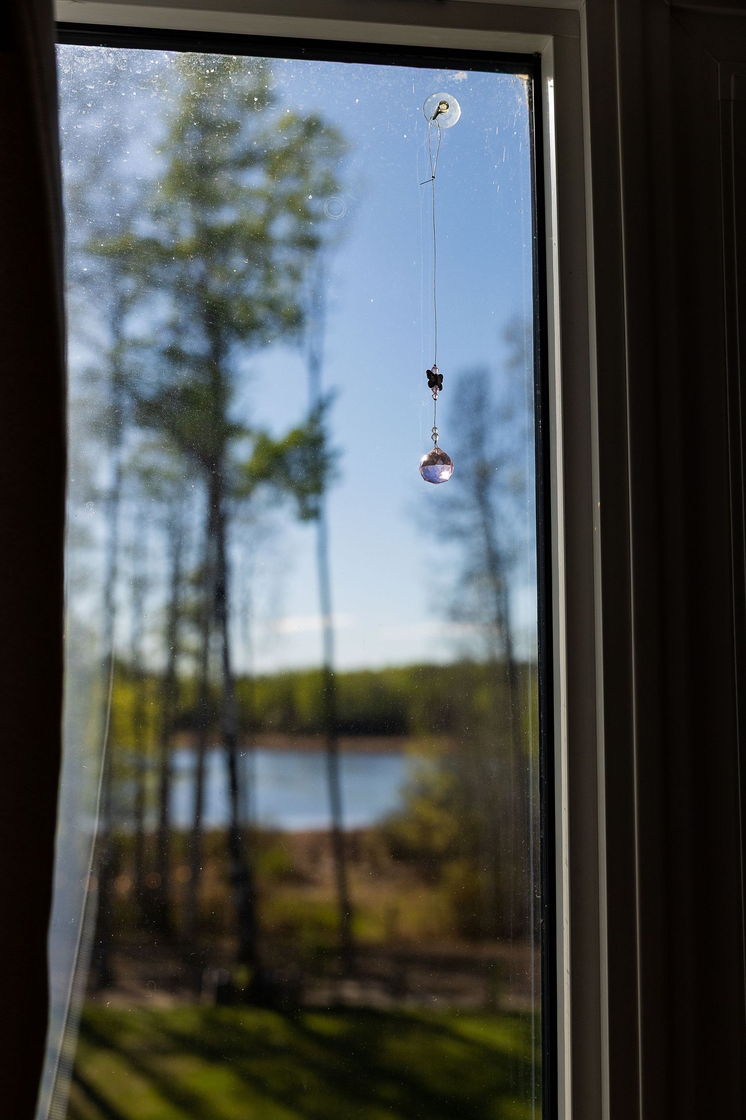 Crystal sun catcher hanging in a window overlooking a serene lake surrounded by trees on a clear day.