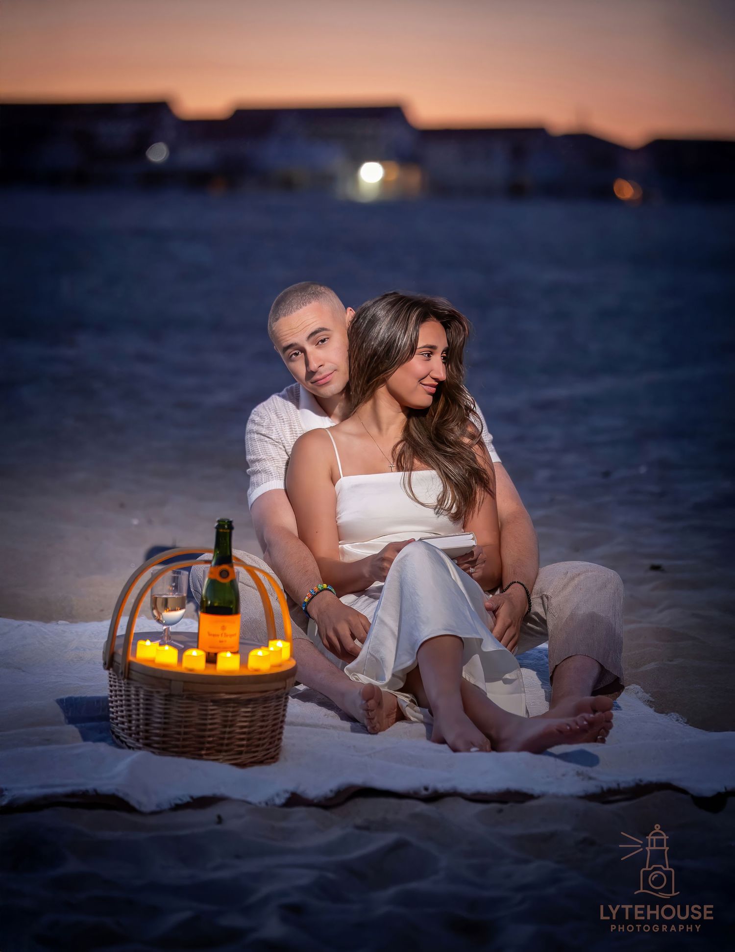 Romantic candlelit beach picnic at sunset with a couple sitting on blanket by glowing lanterns and wicker basket.