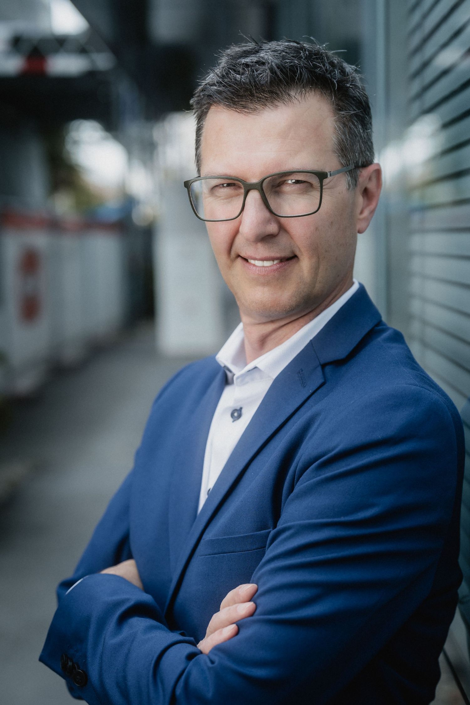 Professional portrait of someone in a navy suit and glasses against an architectural background with blurred elements.