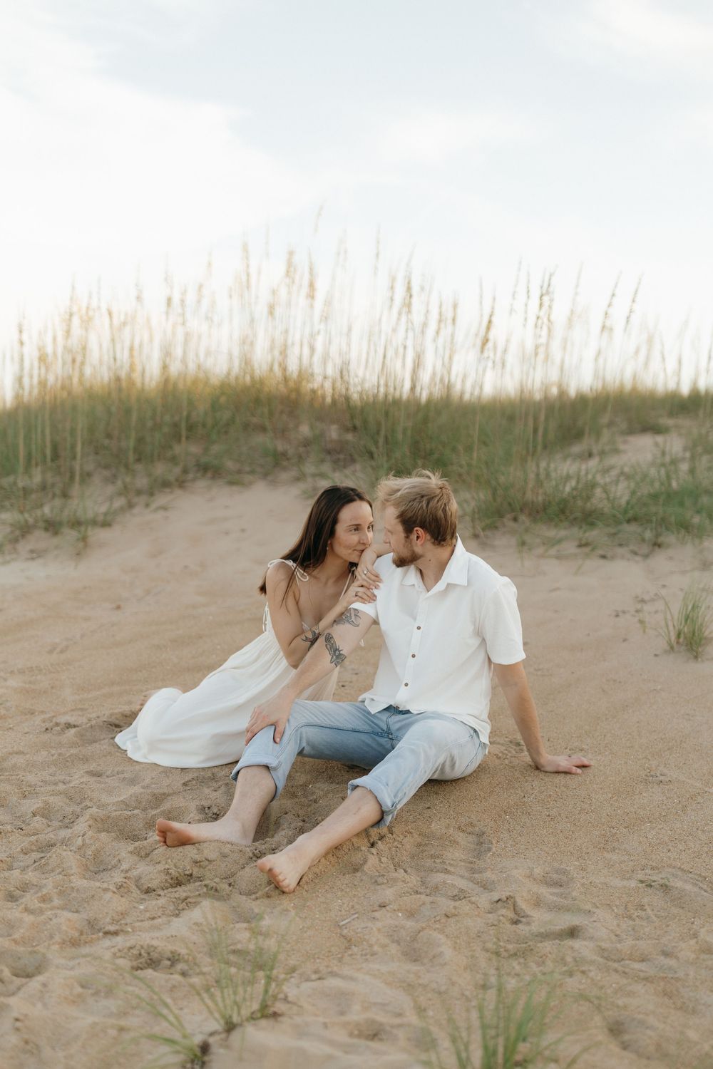 A couple relaxes together on a sandy beach dune with tall grass swaying in the summer breeze.