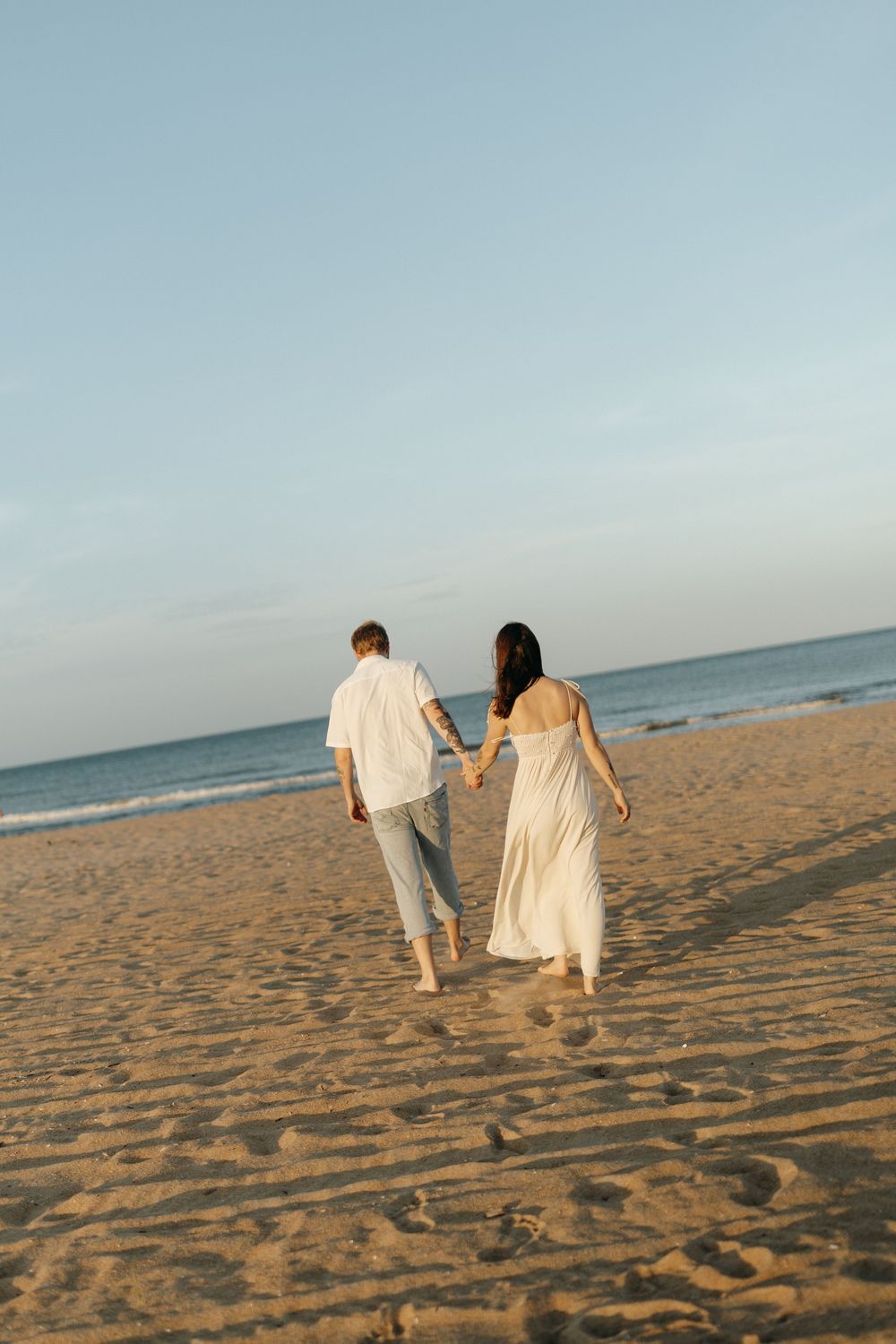A couple in light-colored outfits walks hand in hand along a peaceful sandy beach at sunset.