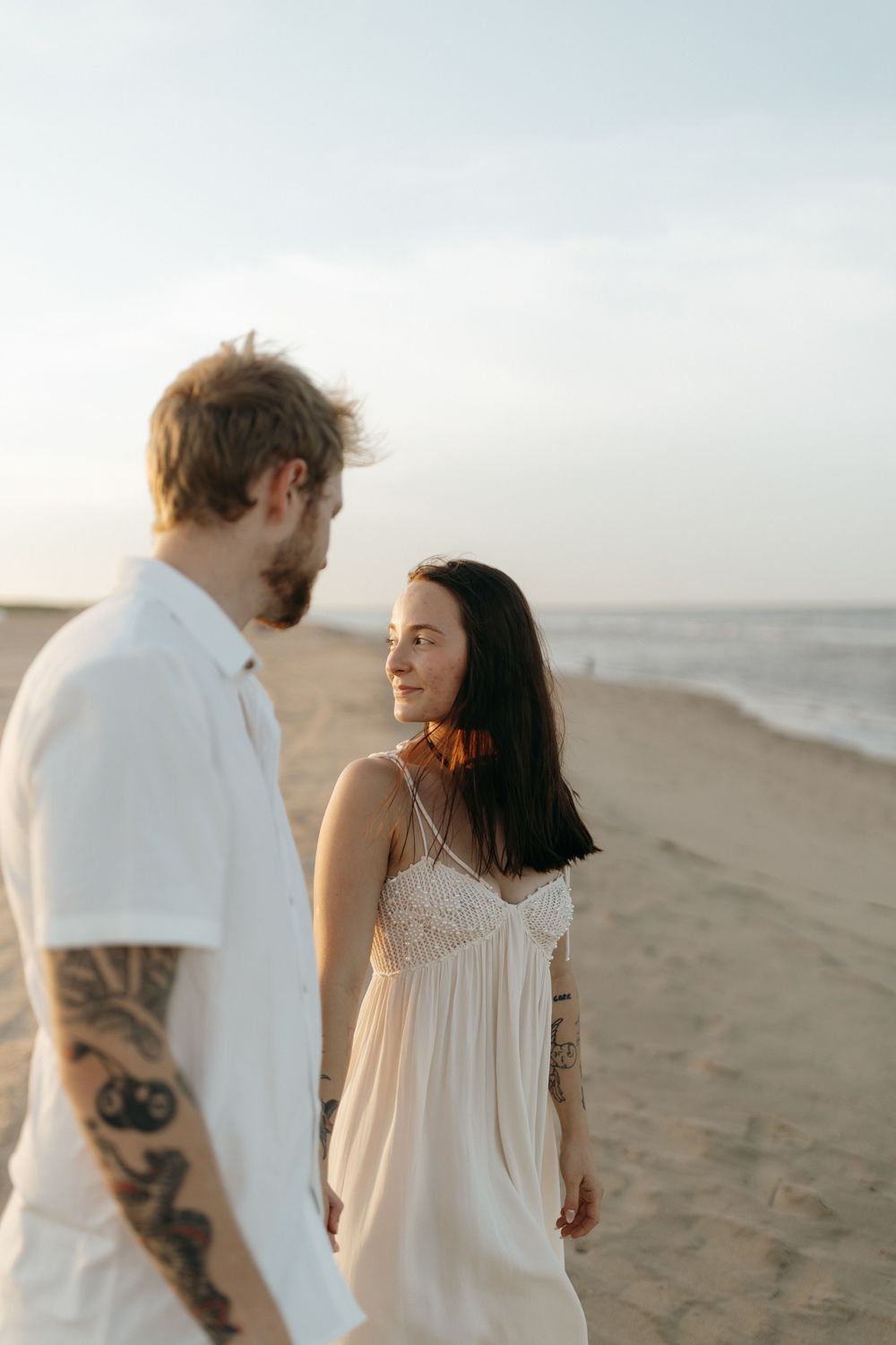 Romantic photoshoot of couple in white outfits walking along a sandy beach at sunset.
