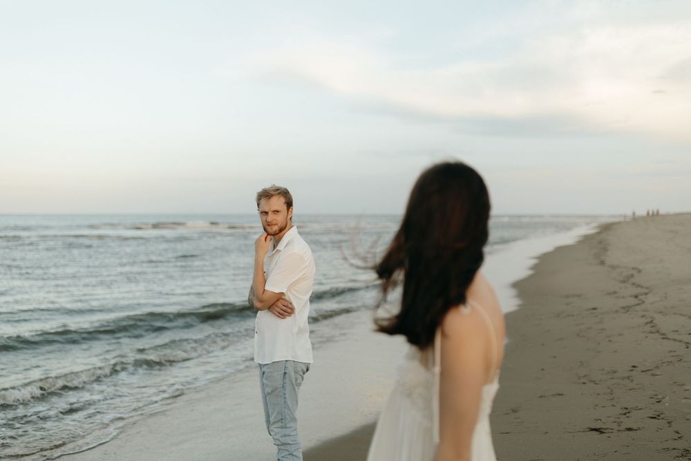 Two people walk along a misty beach at sunset with waves crashing on the shore and cloudy skies above.