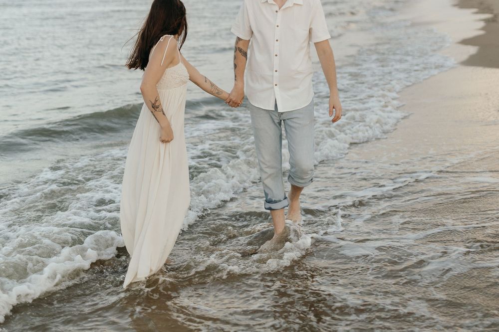 Two people in beachwear hold hands while walking along the shoreline as gentle waves lap at their bare feet.