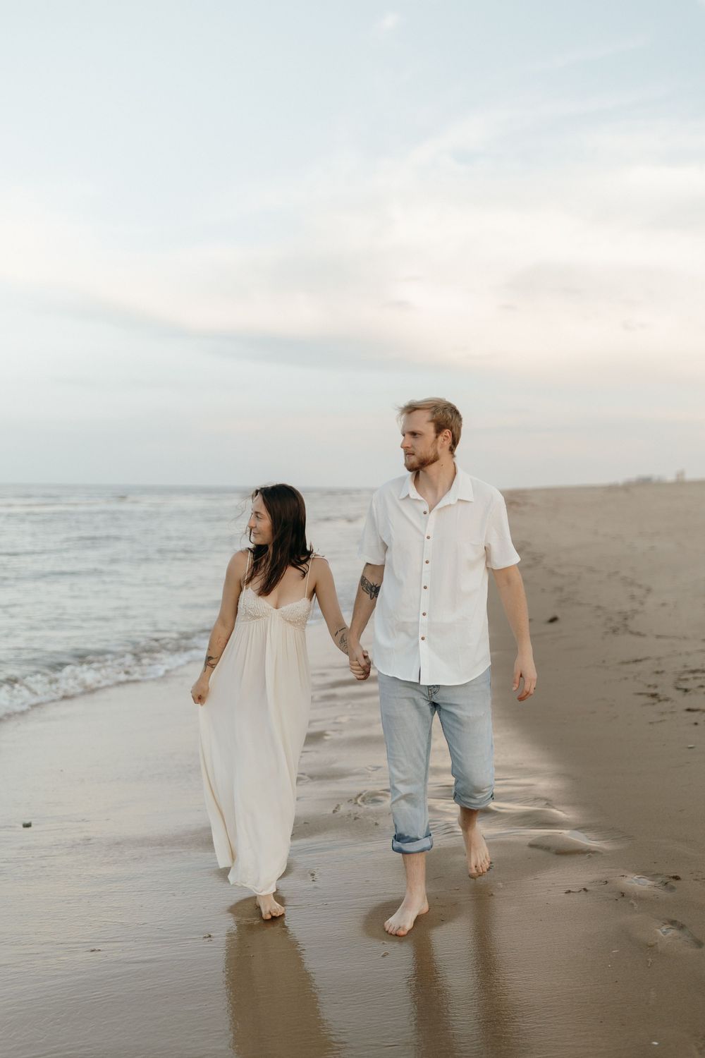 A couple walks hand in hand along a misty ocean beach at sunset wearing white and light colored casual attire.
