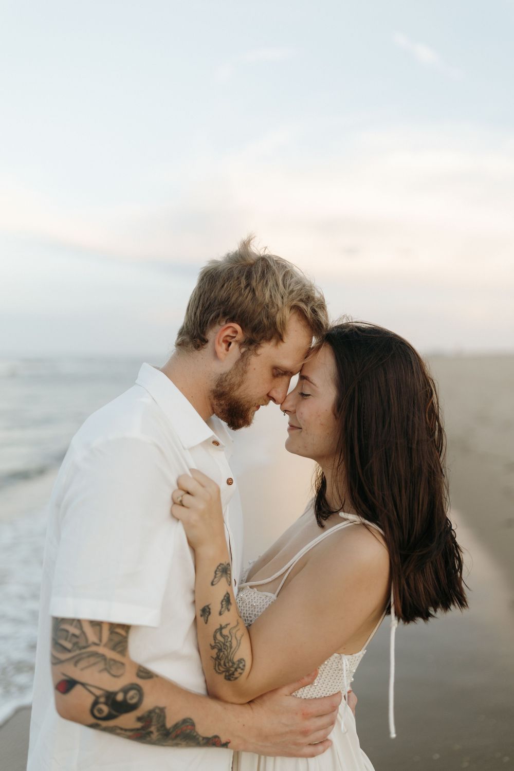 A romantic couple embraces and kisses on the beach at sunset, showcasing their tattoos and white attire.