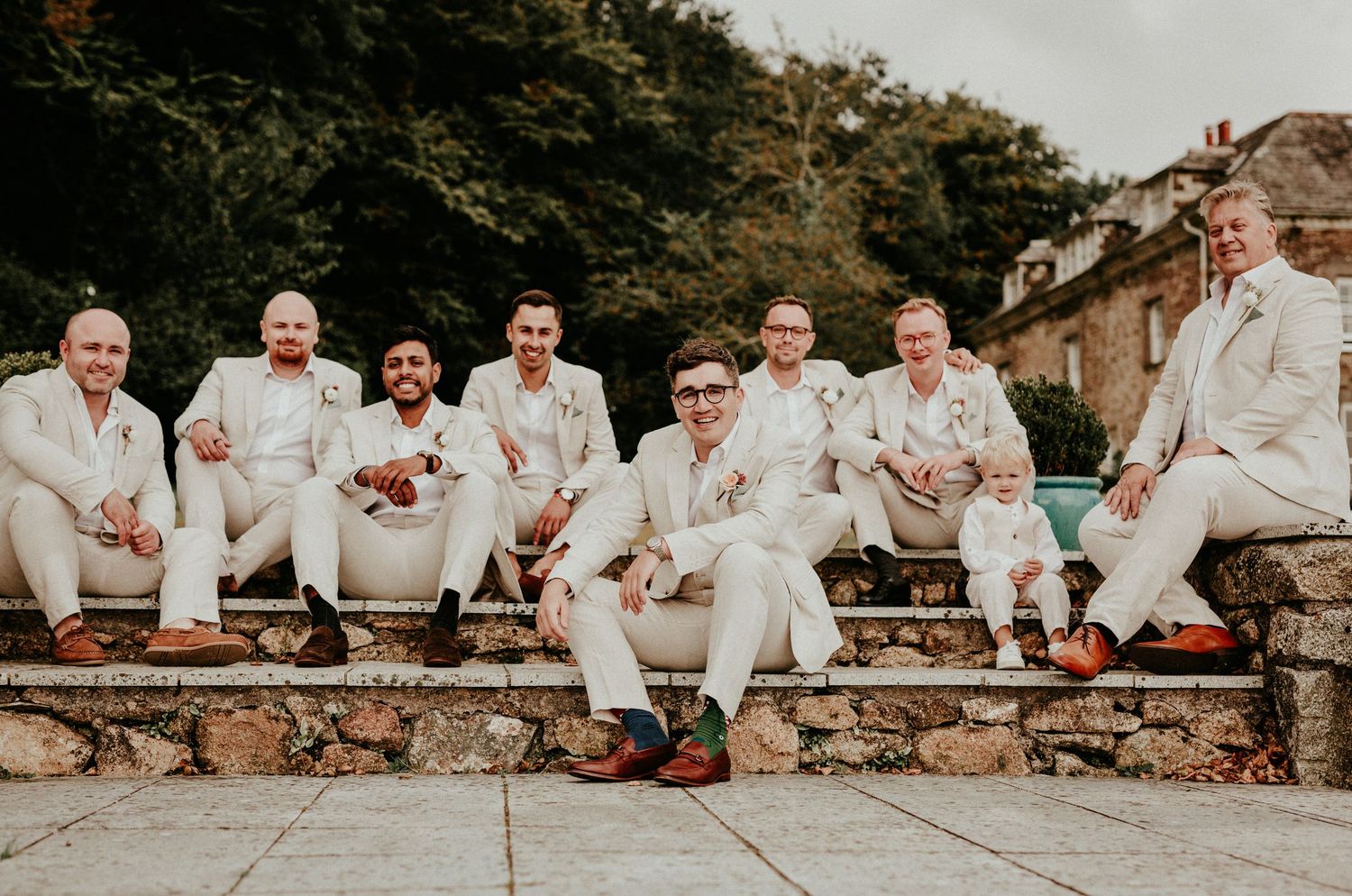 Large group of groomsmen in cream-colored suits pose together on rustic steps at an outdoor wedding venue.