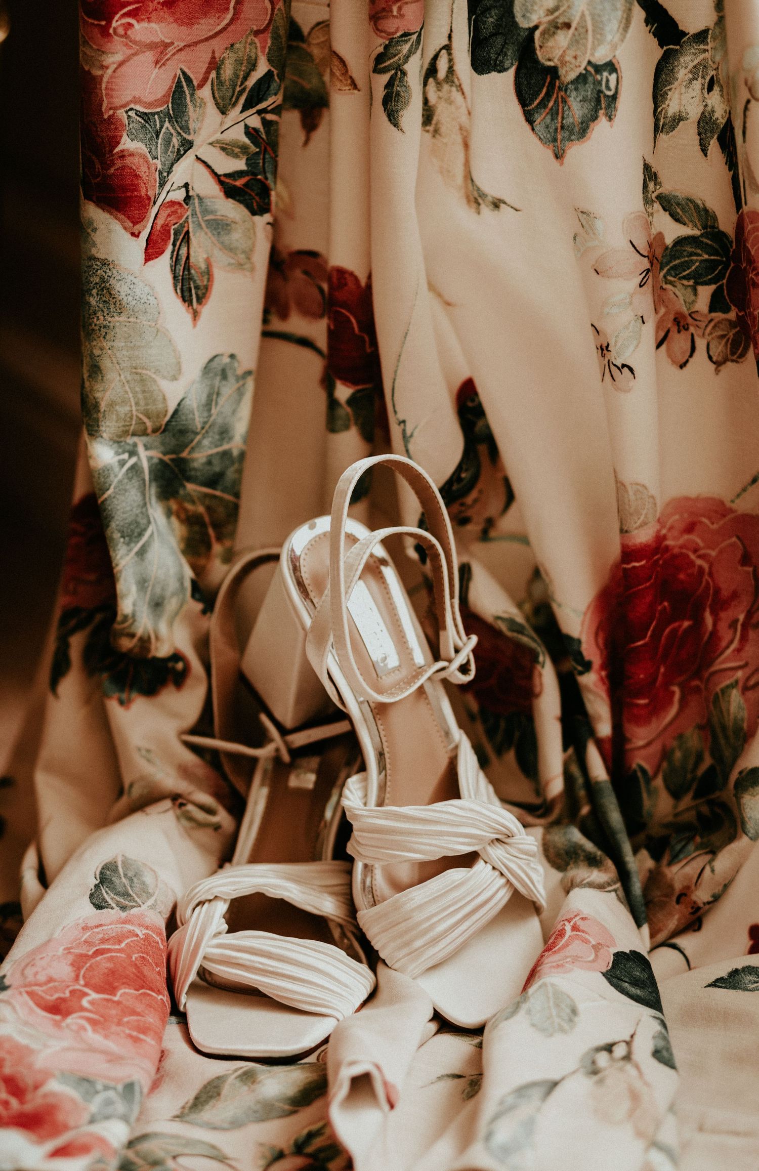 Elegant white strappy wedding sandals displayed against a floral patterned backdrop in soft lighting.
