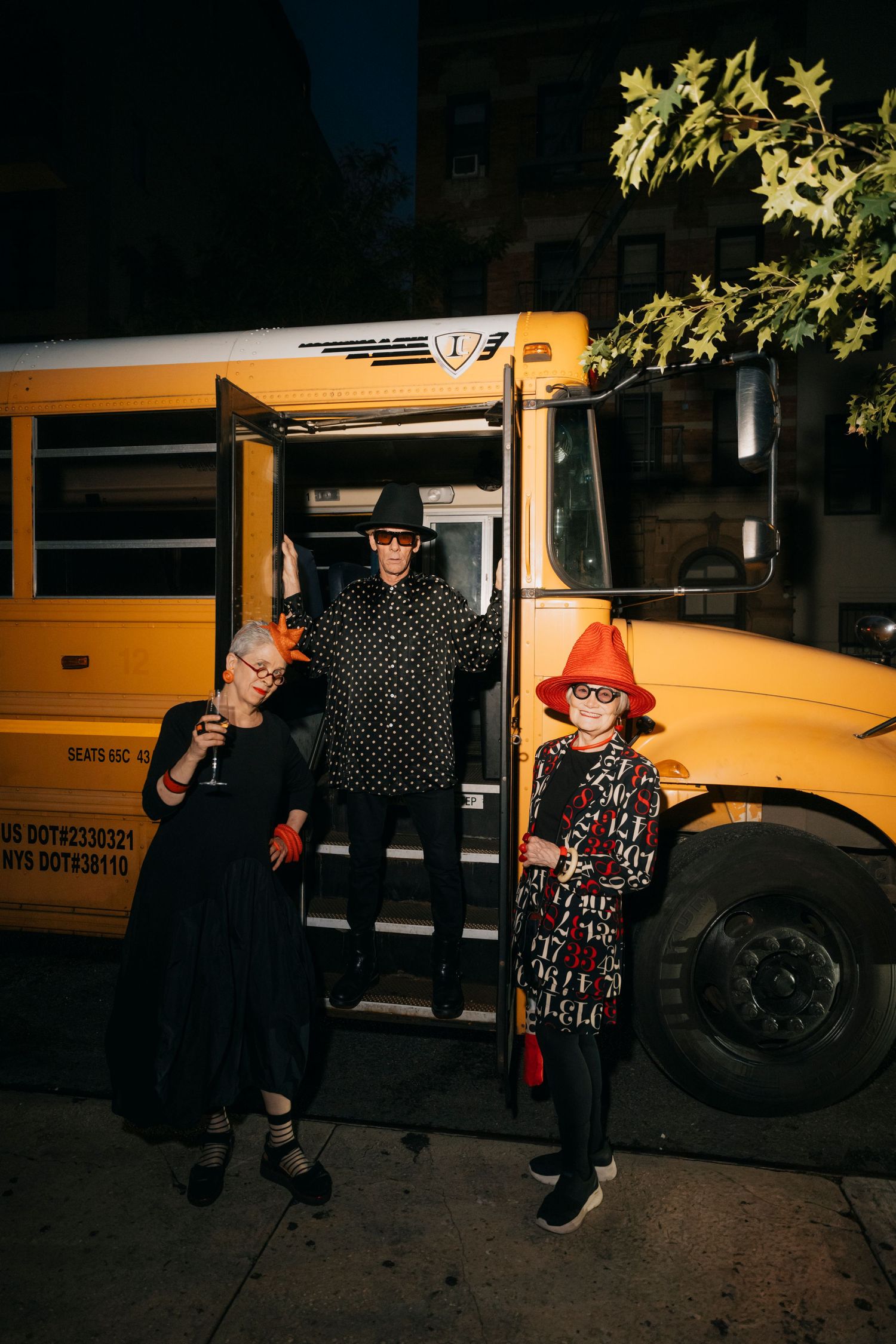Three stylishly dressed people pose in front of a yellow school bus at night with tree branches visible overhead.