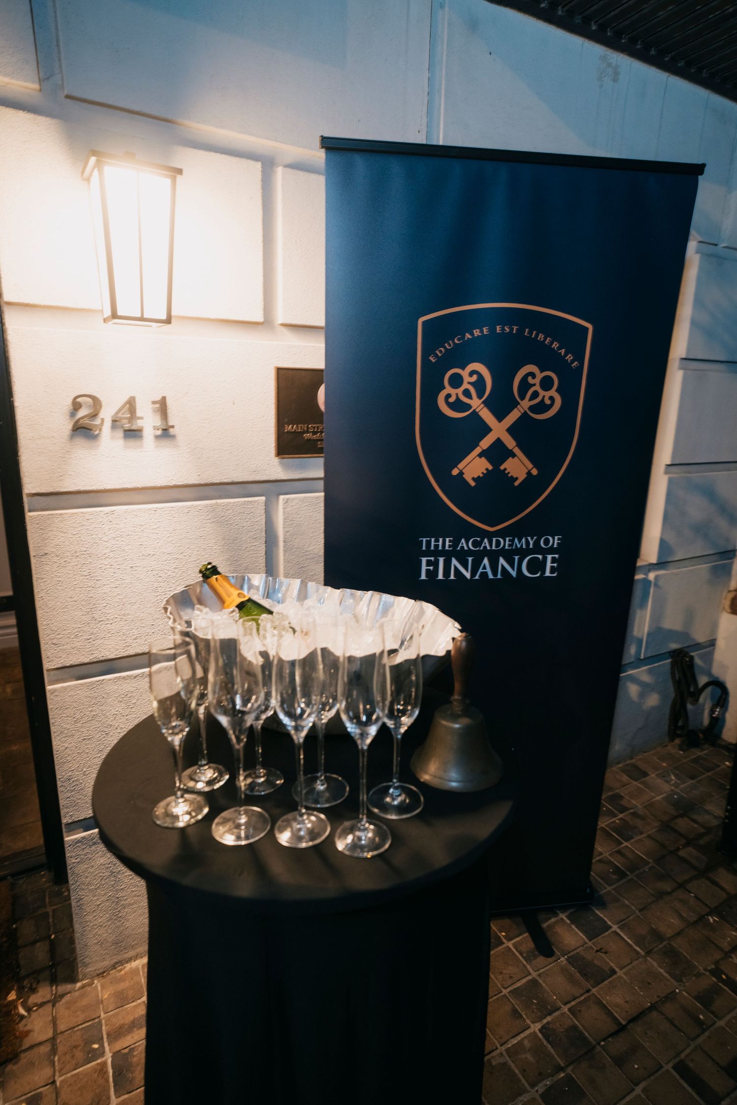 Glass champagne flutes arranged in front of black branded backdrop with institutional logos and elegant lighting.