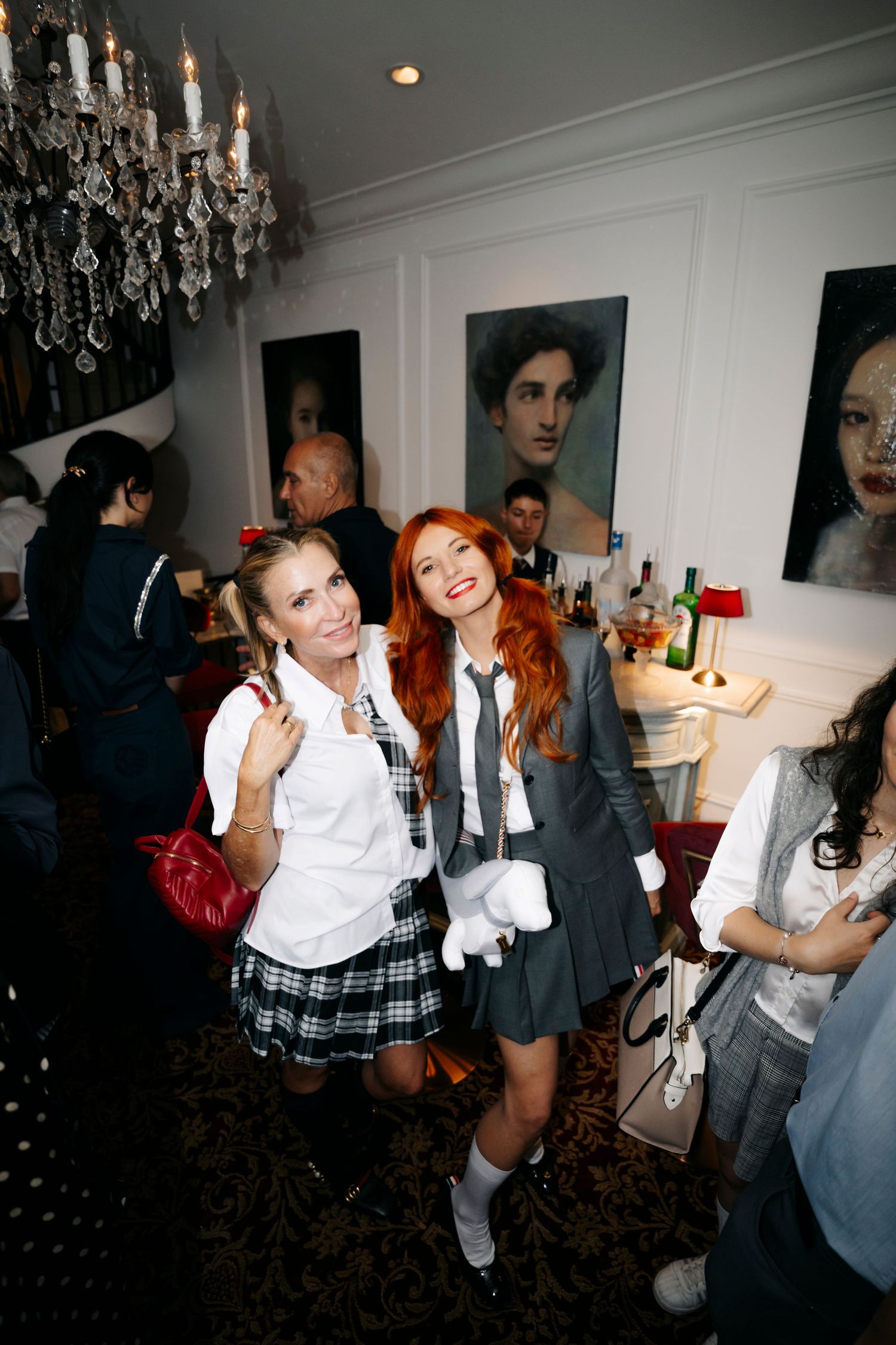 Two students in school uniforms pose together at a party with portrait artwork and a crystal chandelier in the background.