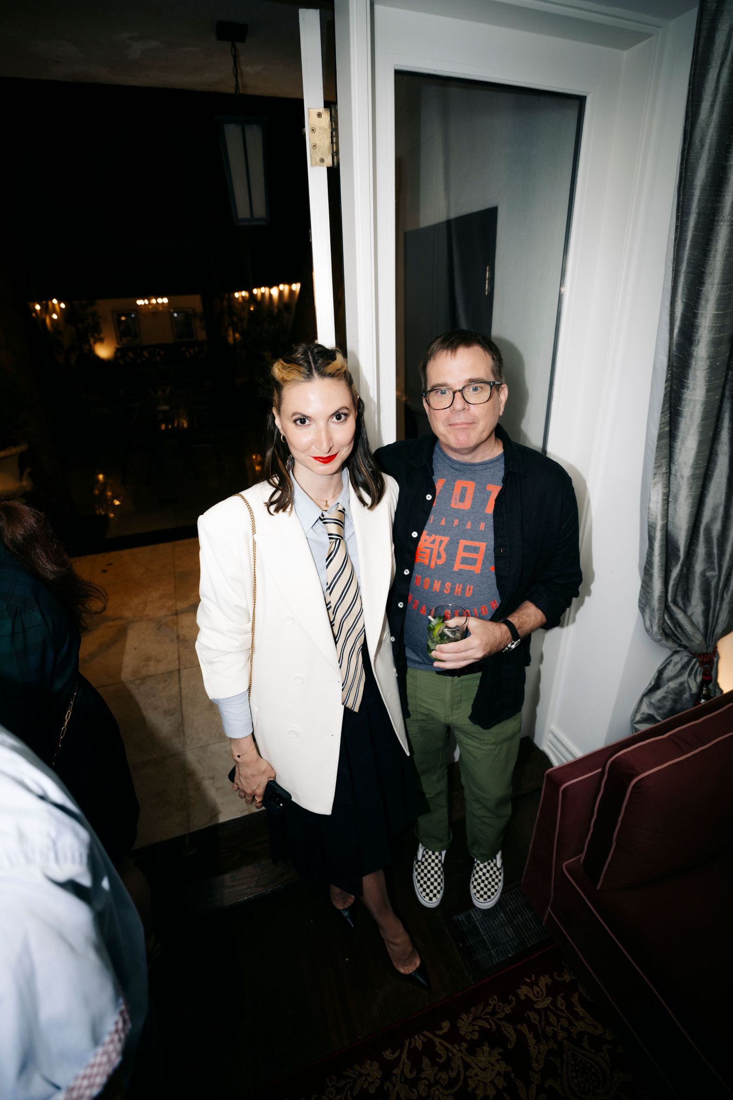 Two people posing together at a party, one wearing a white blazer with tie and another in a graphic t-shirt.