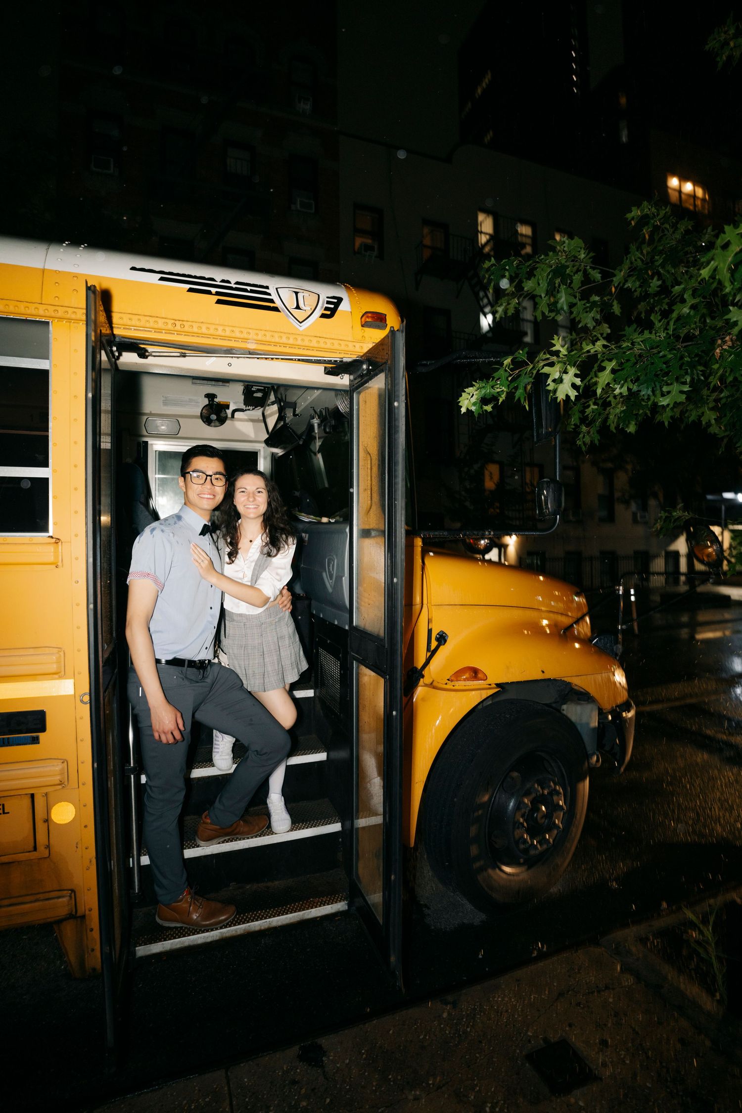 A couple poses in the doorway of a yellow school bus at night on a city street.