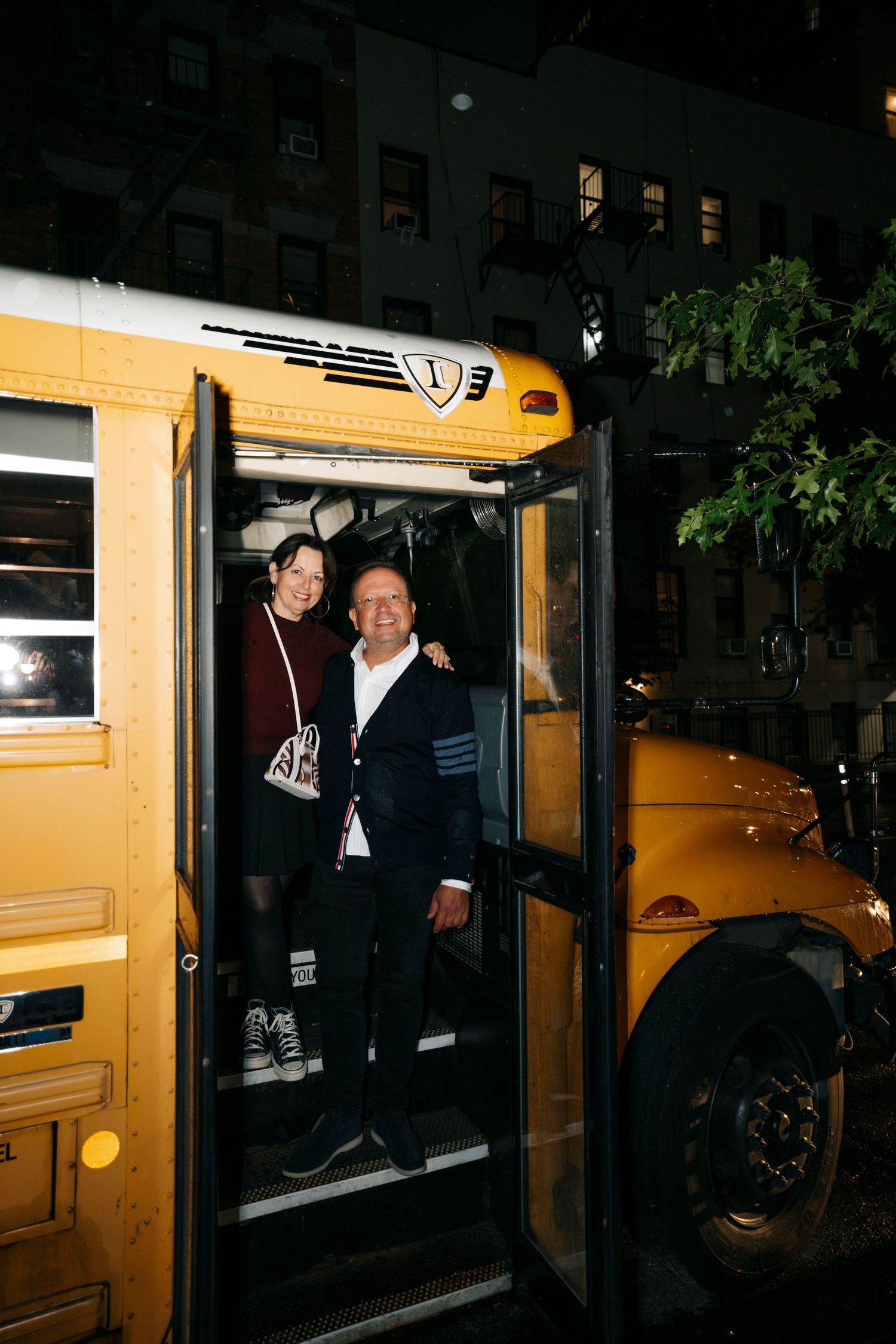Two people stand in the doorway of a yellow school bus at night in an urban setting.
