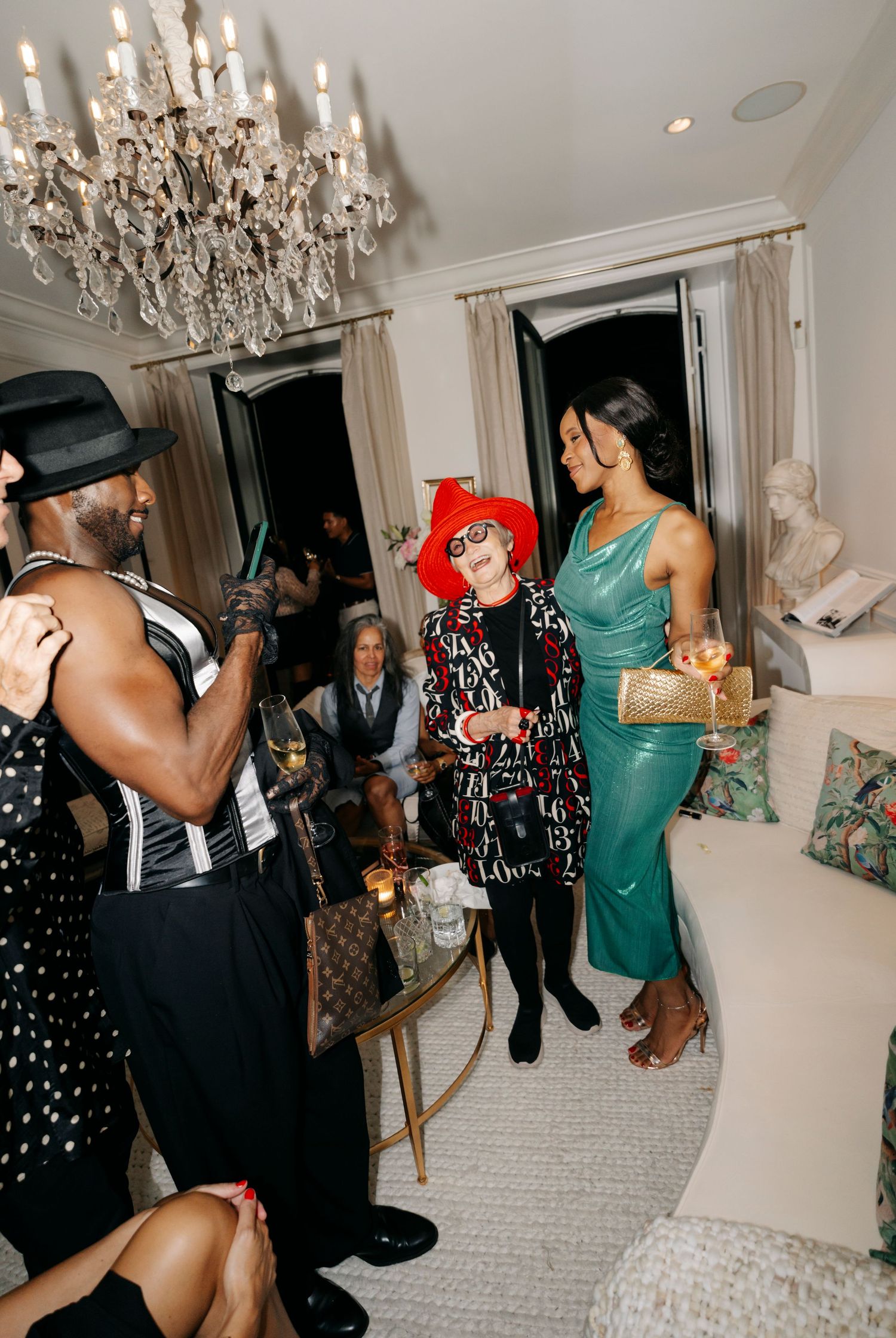 Partygoers dressed in elegant outfits socializing under a crystal chandelier in a luxurious white room.