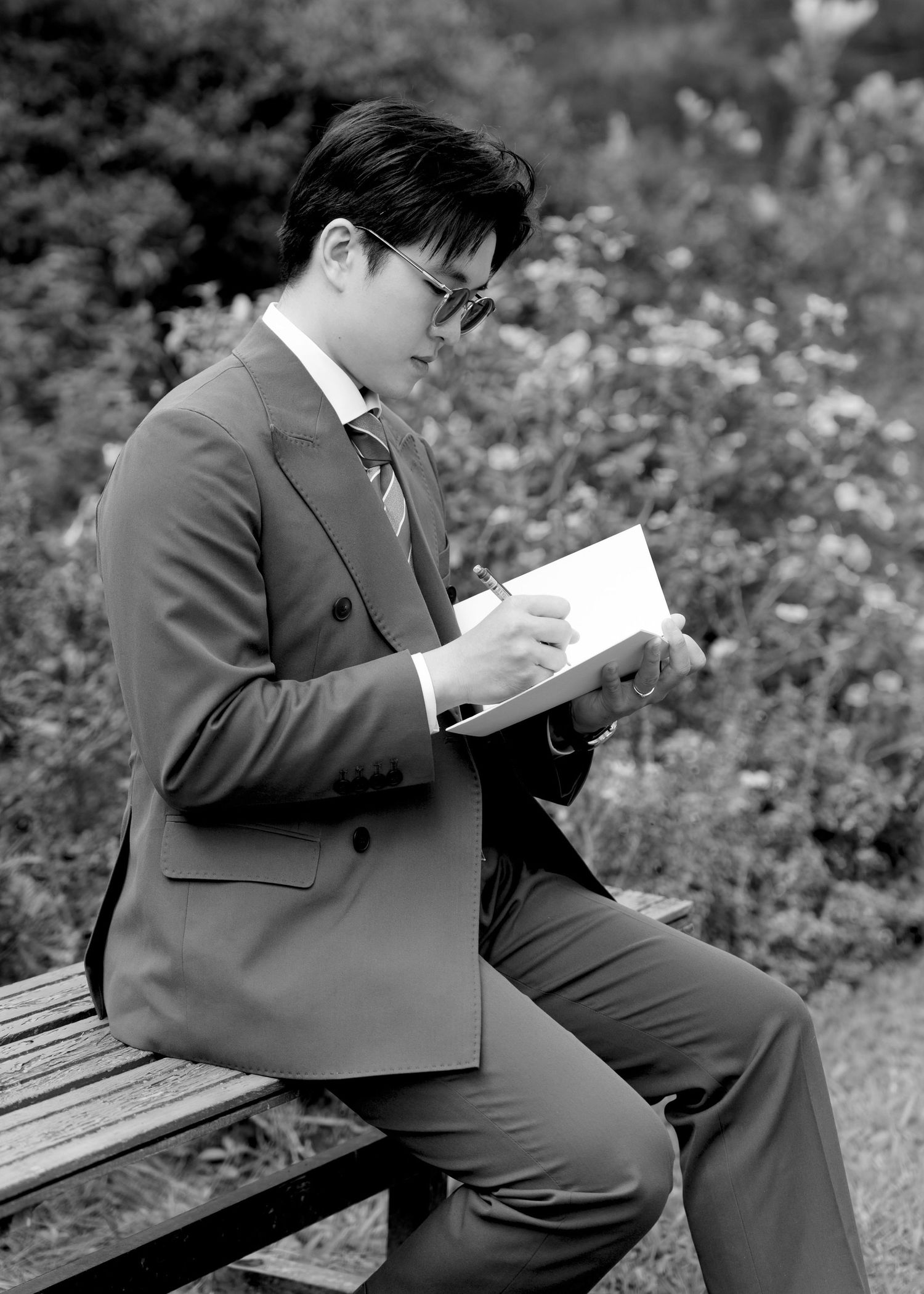 Black and white photo of man in suit writing in notebook while seated on park bench outdoors.