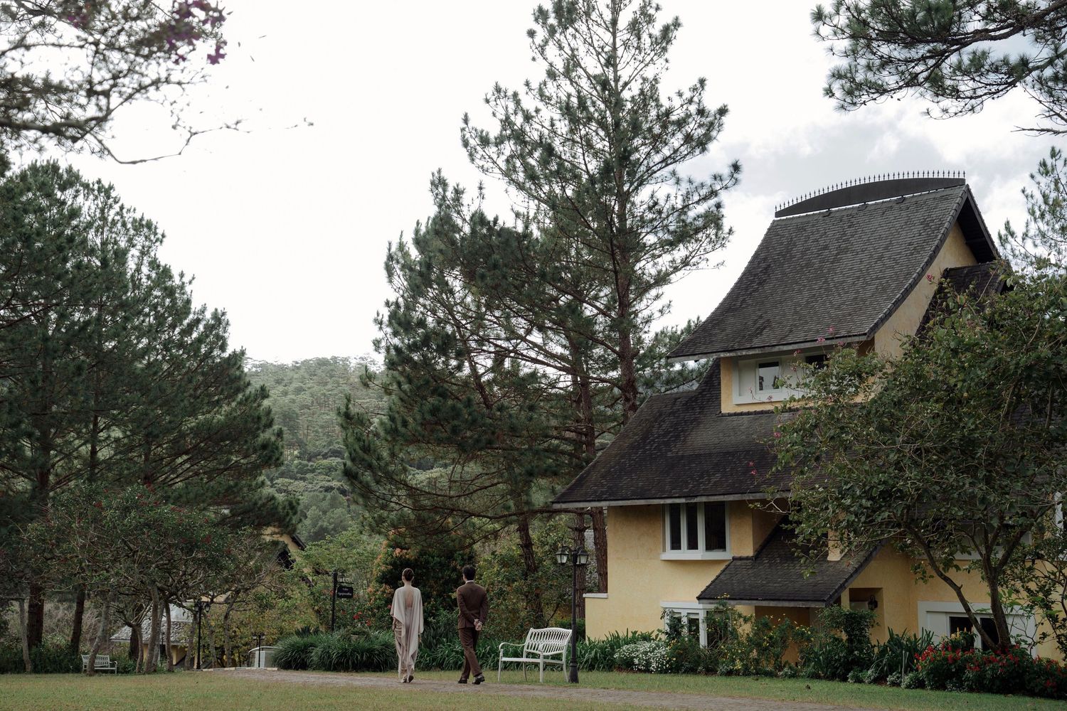 A couple walks near a charming yellow villa surrounded by tall pine trees in a scenic garden setting.