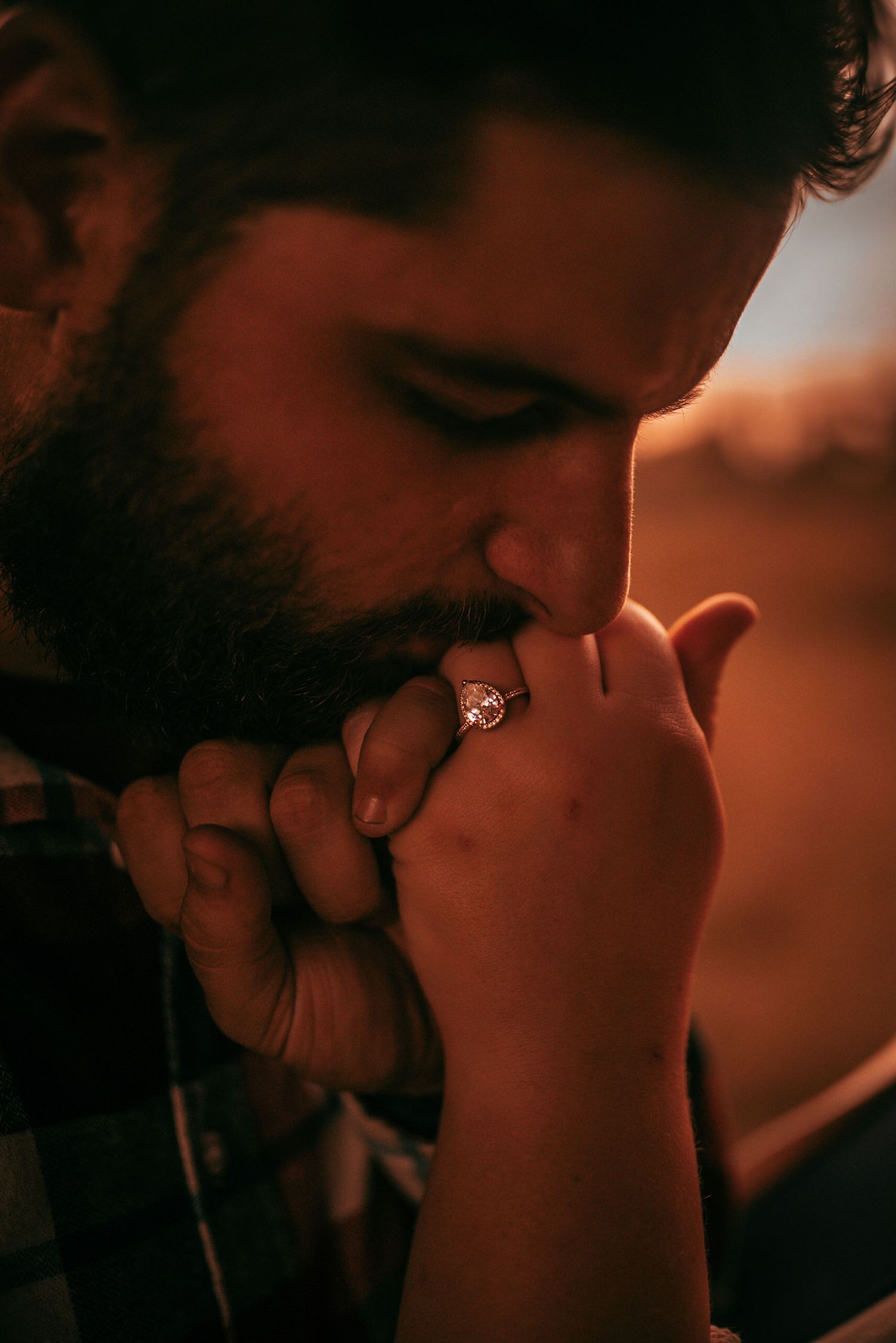 An intimate engagement photo showing a close-up of a ring kiss at sunset.