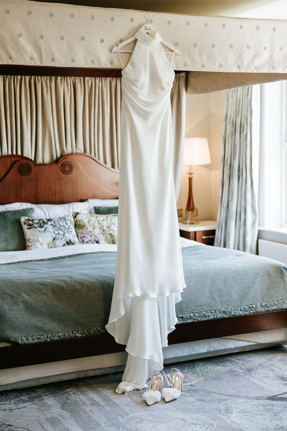 Elegant white wedding dress with halter neckline and flowing train hanging above a bed with sage green bedding and silver shoes.