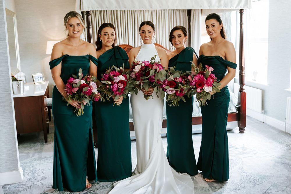 Bridesmaids in emerald green dresses holding pink floral bouquets pose alongside bride in elegant white gown.