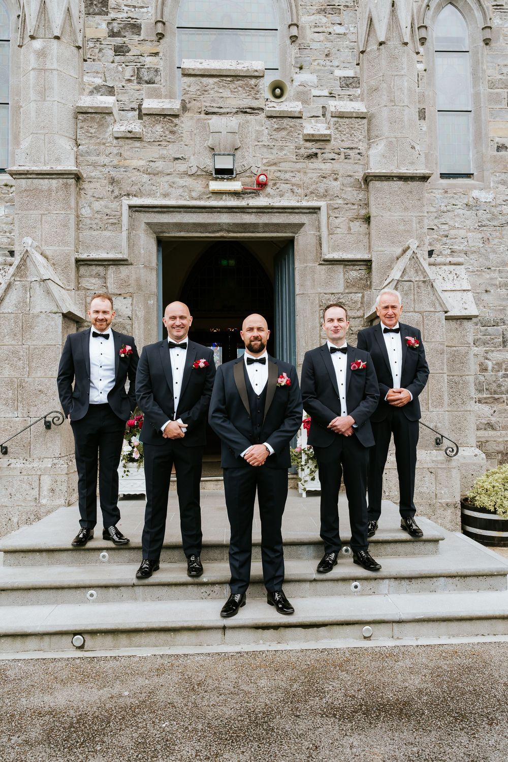 Groomsmen in black tuxedos stand lined up on stone steps outside a historic church building.