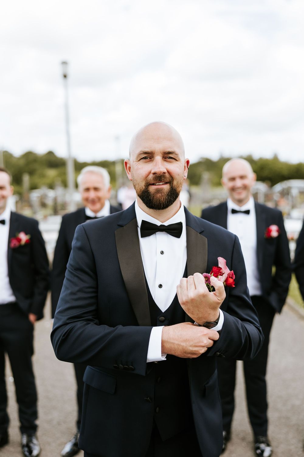 Wedding party member in black tuxedo with bow tie and red boutonniere adjusts his cuff links outdoors.