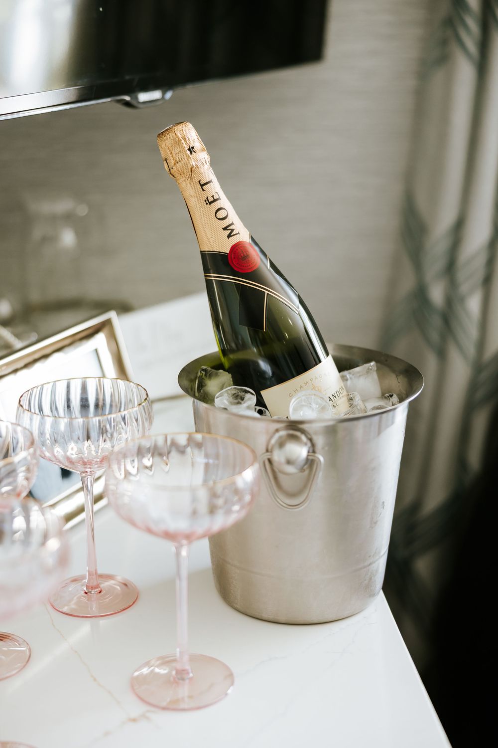 Elegant champagne bottle chilling in a silver ice bucket with pink-tinted coupe glasses arranged on a white surface.