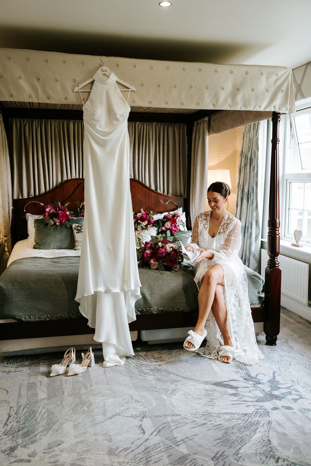 Wedding dress hangs elegantly in a luxurious hotel room with bouquet and bridal accessories laid out on bed.