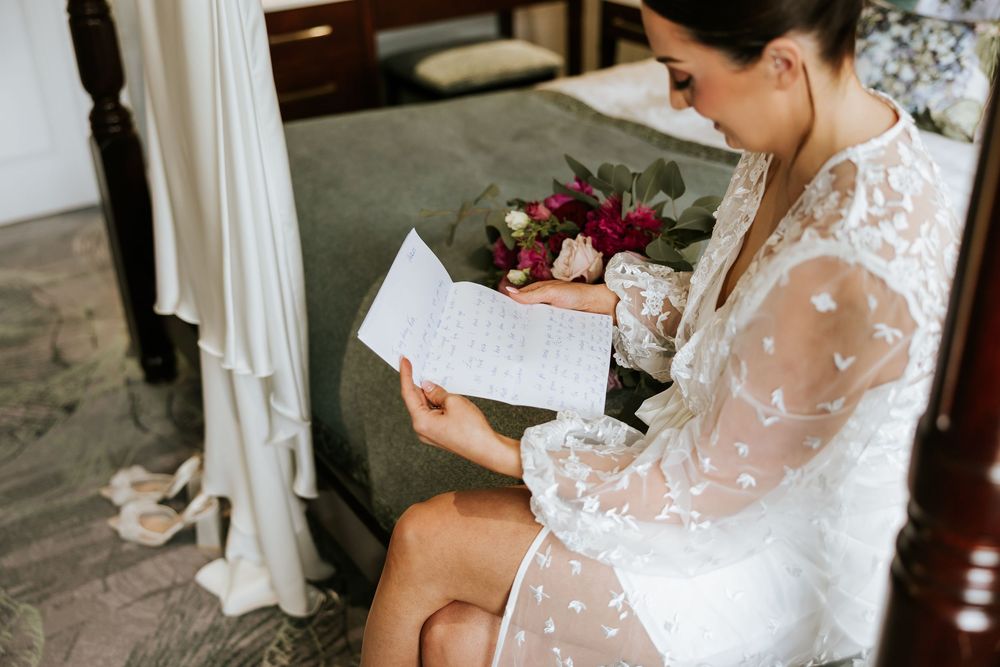 A bride in a delicate lace wedding dress sits elegantly while reading a note before her ceremony.