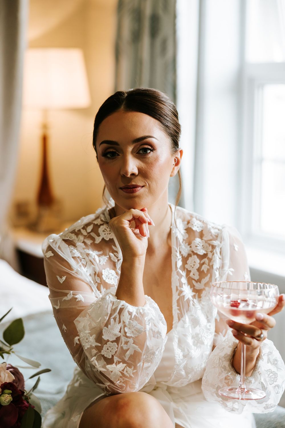 A woman in a white lace wedding dress holding a champagne glass near a window with warm lighting.