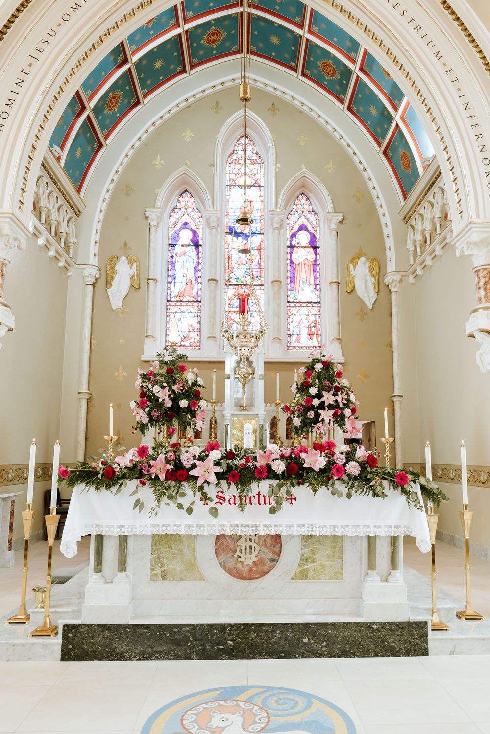 Beautiful church altar decorated with pink and white floral arrangements and stained glass windows in gothic arched design.
