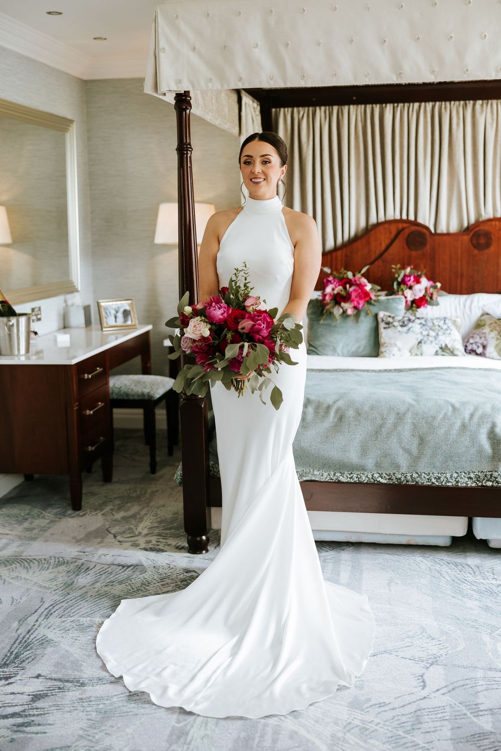 A beautiful bridal suite featuring a white wedding dress draped elegantly against a luxurious four-poster bed and floral decor.