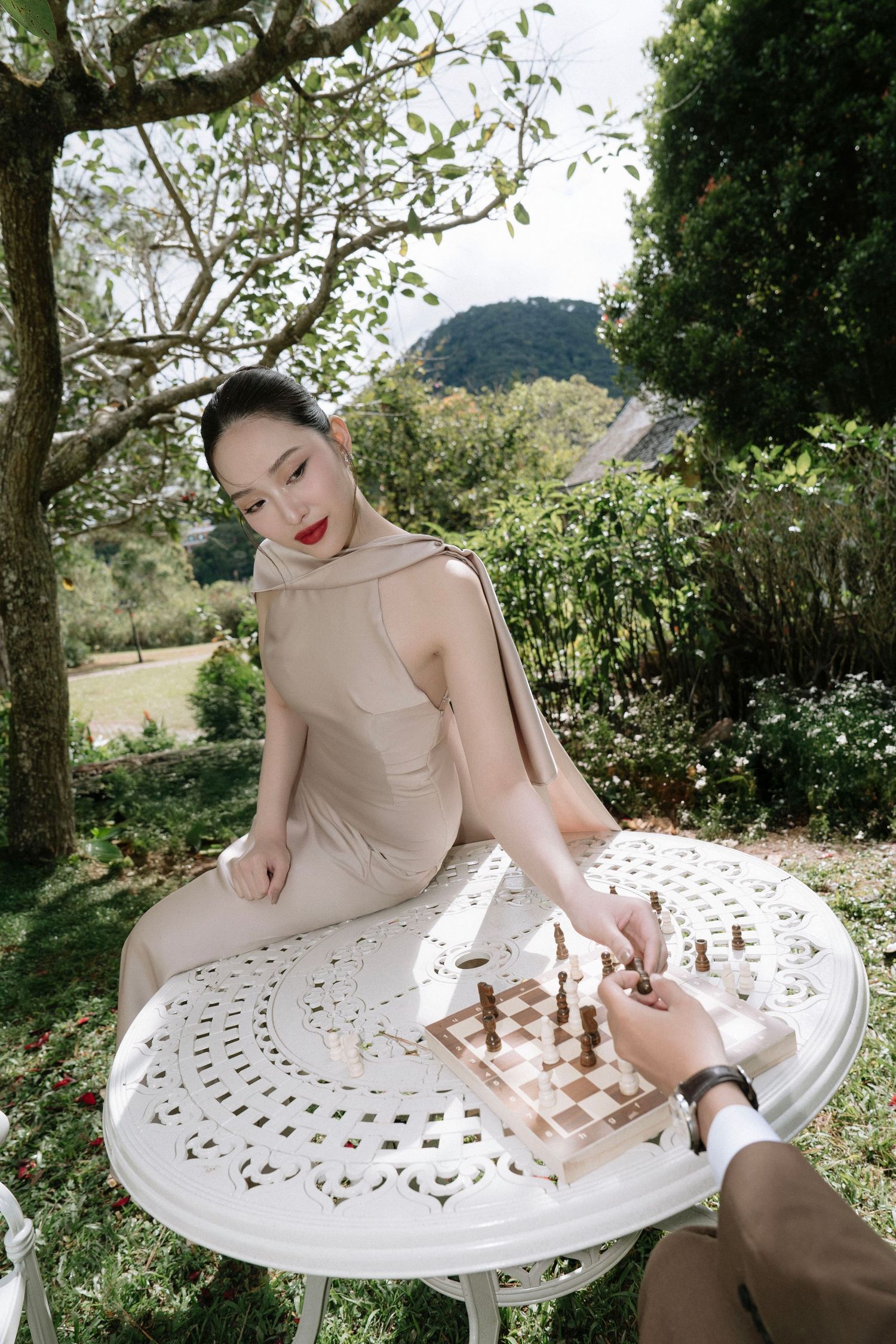 Beautiful woman in beige halter dress playing chess at white garden table in scenic outdoor setting.
