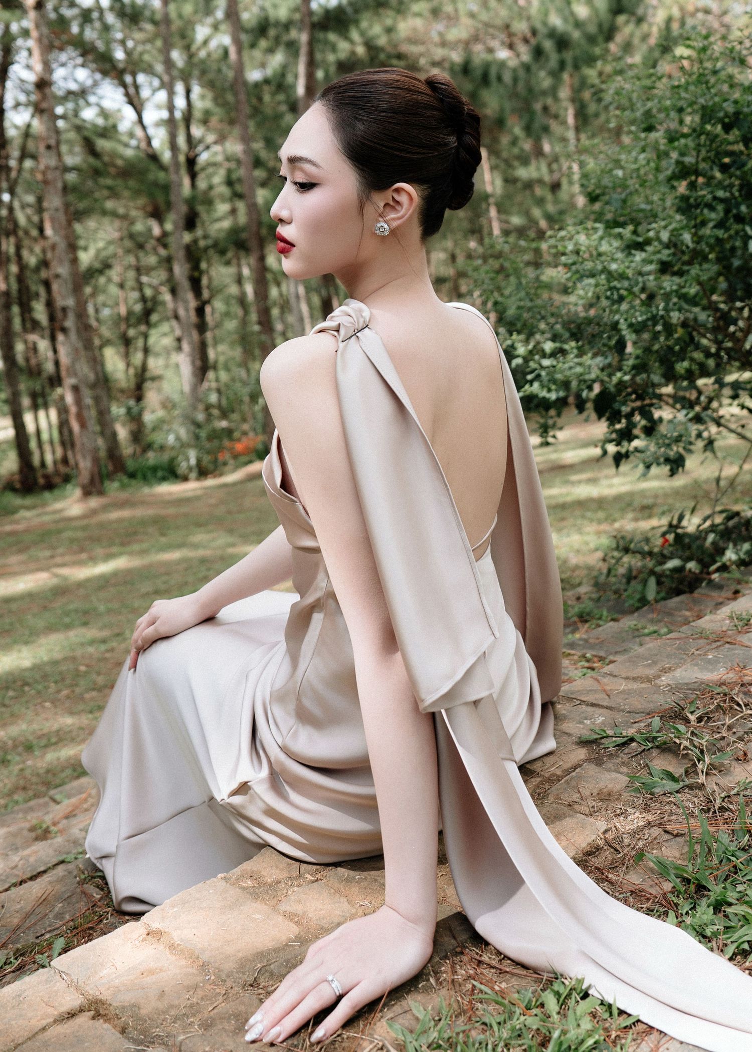 Elegant woman in backless champagne satin gown seated outdoors among pine trees.