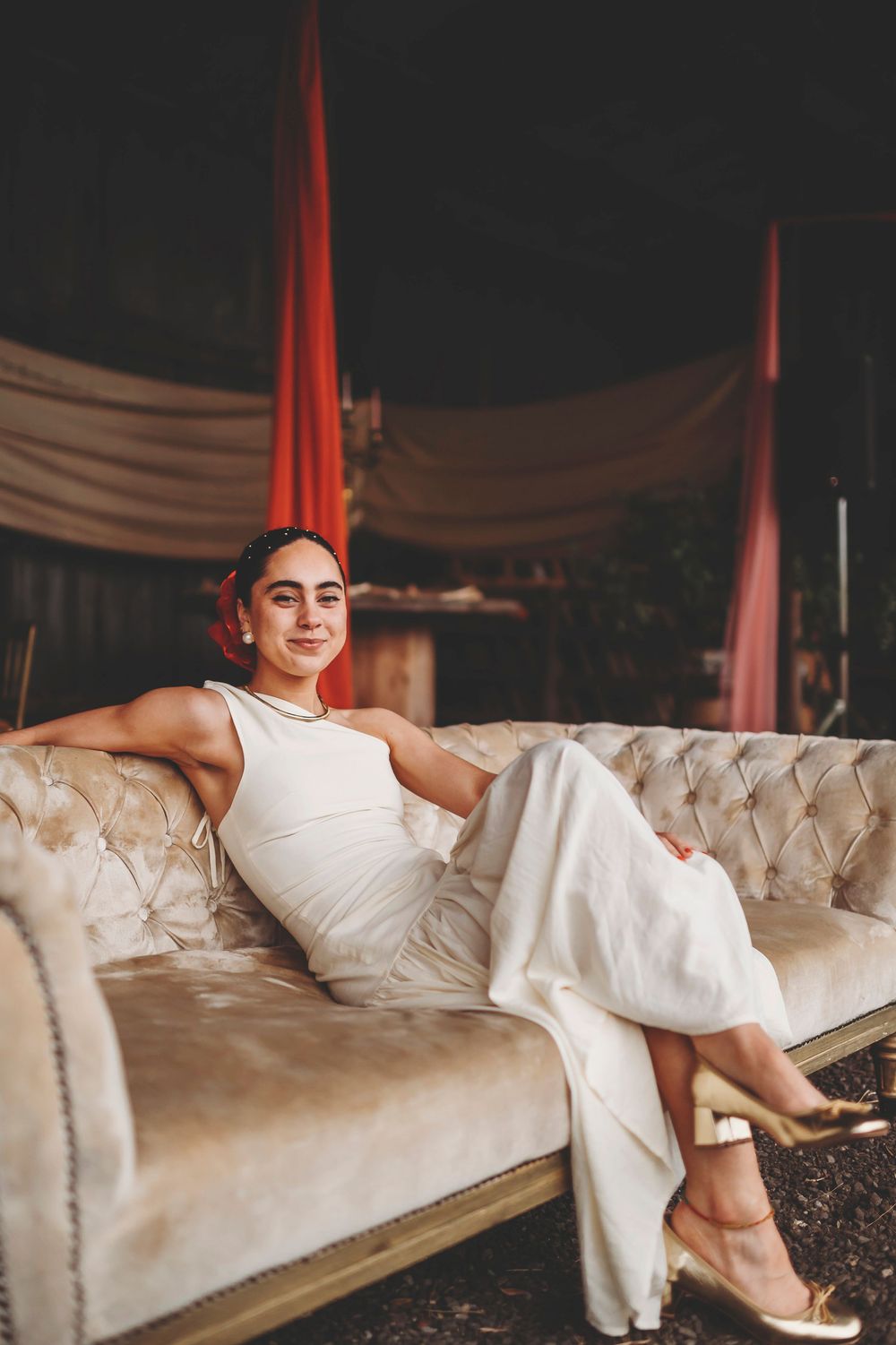 Person in white dress lounges elegantly on plush cream-colored sofa in moody romantic setting.