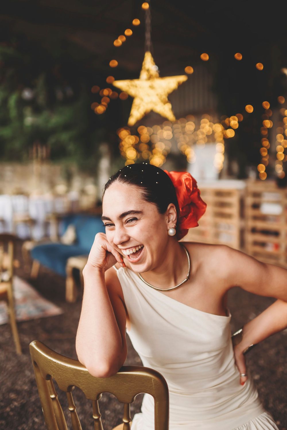 Person in white dress seated on wooden chair laughs joyfully with holiday star lights twinkling in background.