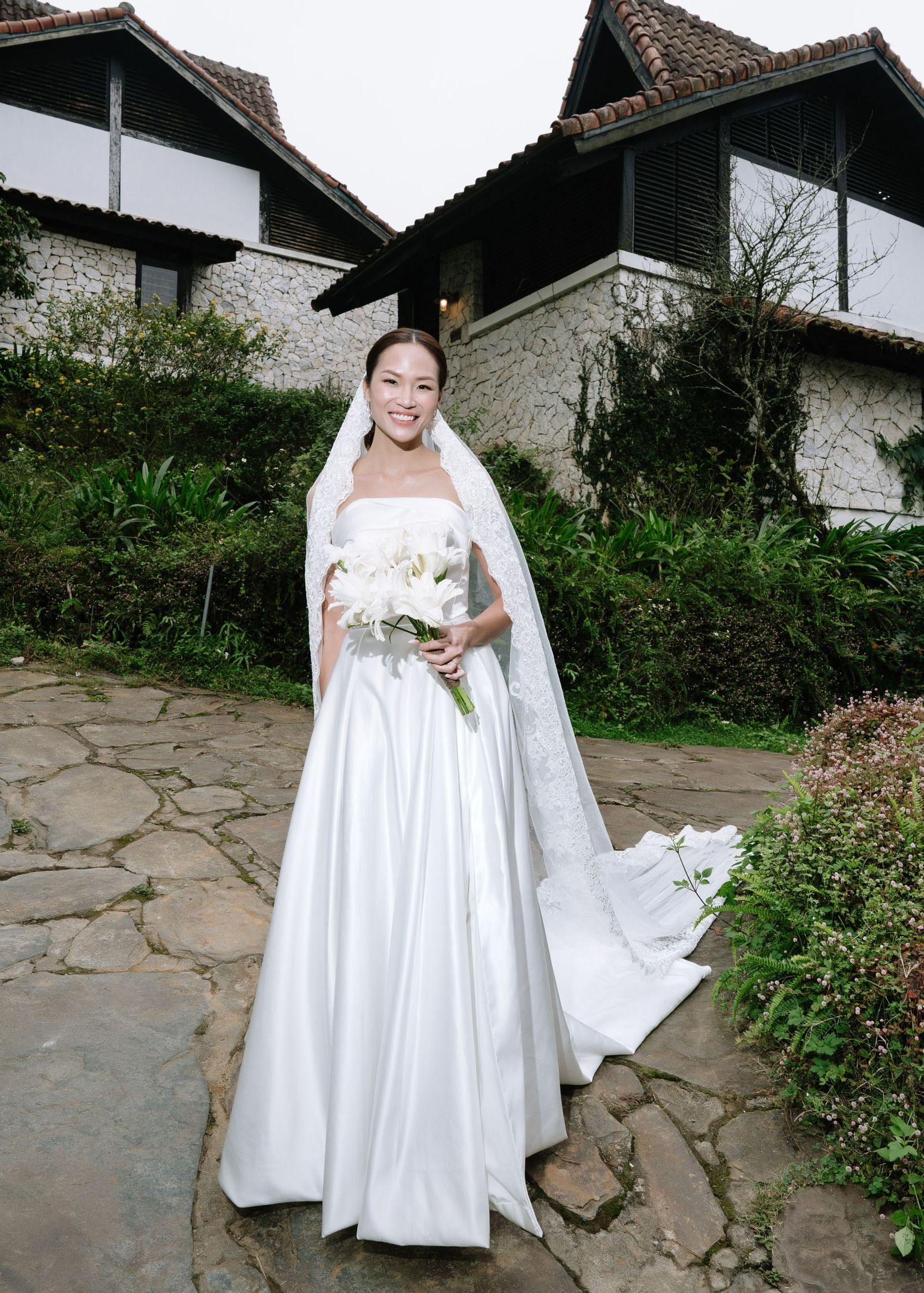 Smiling bride in white strapless gown with veil holding white bouquet outdoors.